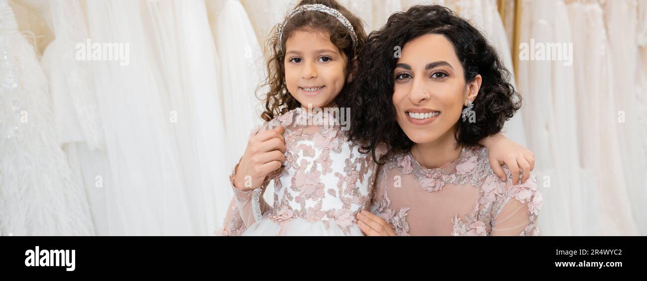 special moment, cheerful middle eastern bride in floral wedding gown sitting and embracing her little daughter in bridal salon around white tulle fabr Stock Photo