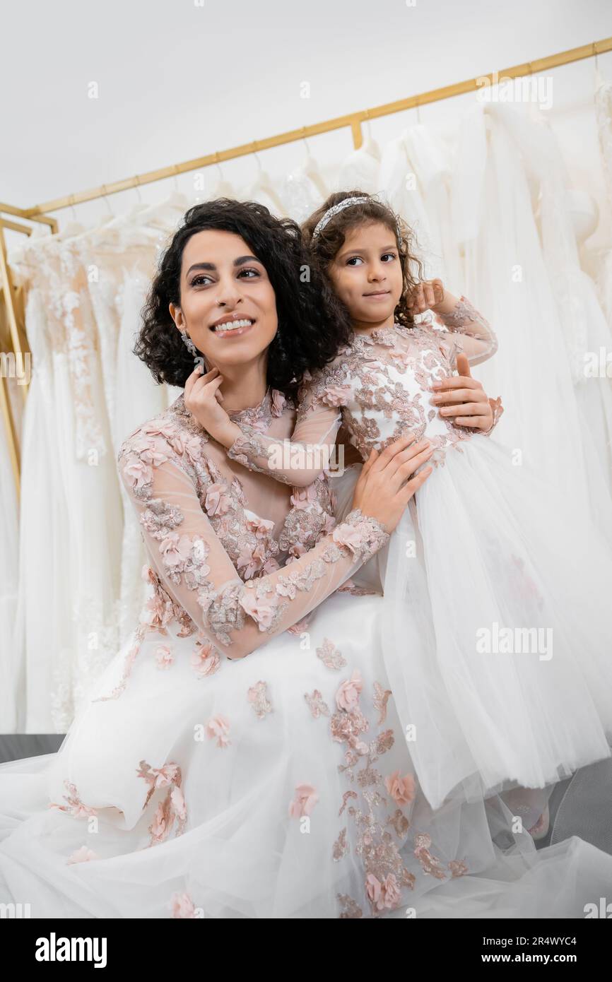 special moment, delightful middle eastern bride in floral wedding gown hugging her little daughter in bridal salon around white tulle fabrics, bridal Stock Photo