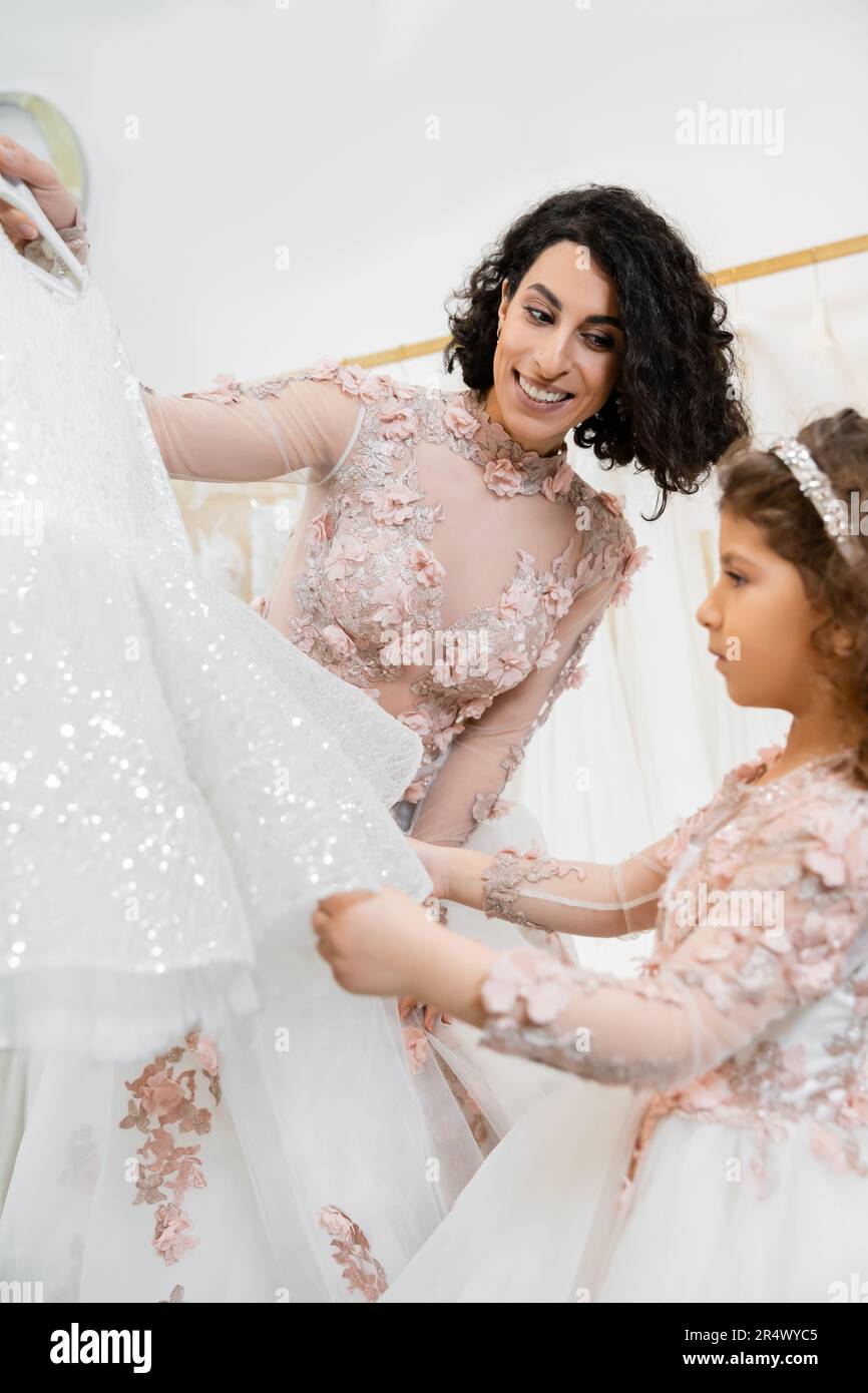 charming and brunette middle eastern woman in floral wedding gown helping to choose dress for her little daughter in bridal salon around white tulle f Stock Photo