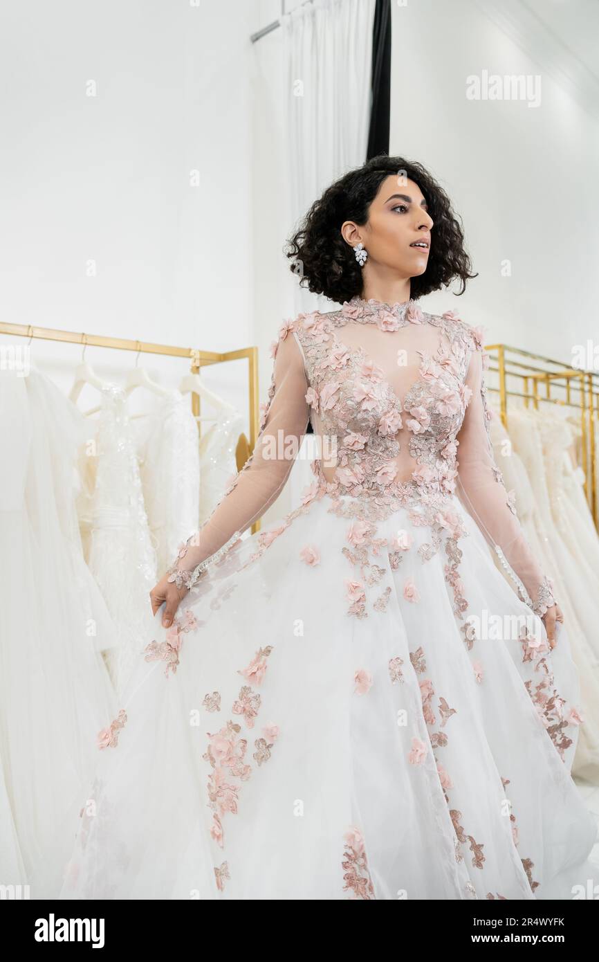 enchanting and middle eastern woman trying on gorgeous and floral wedding dress inside of luxurious bridal salon, shopping, bride-to-be,  blurred whit Stock Photo