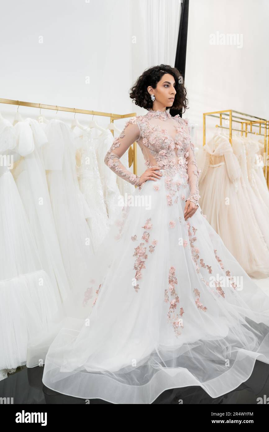 brunette and middle eastern woman with wavy hair trying on gorgeous and floral wedding dress with train inside of luxurious bridal salon, shopping, br Stock Photo