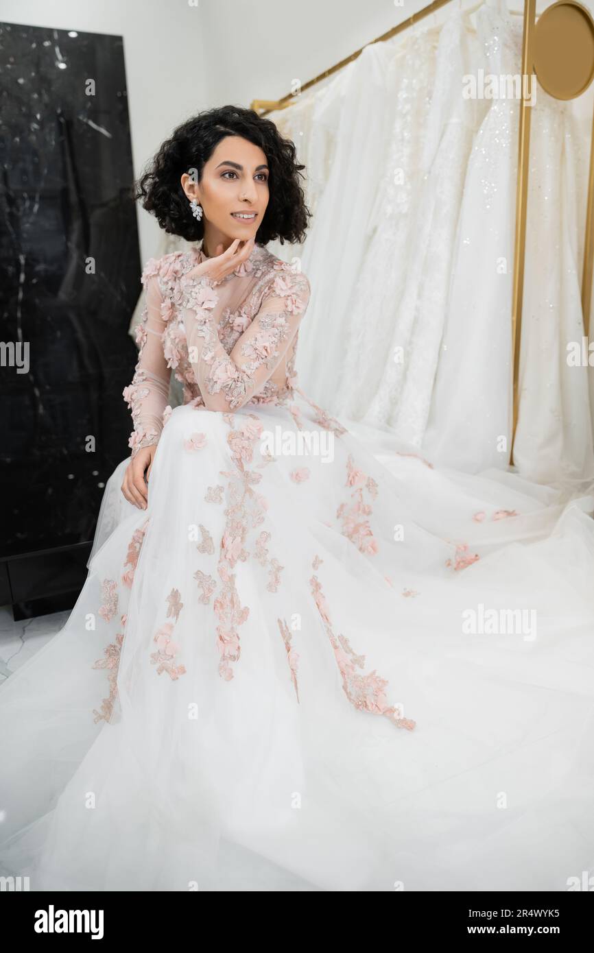 happy middle eastern woman with wavy hair sitting in gorgeous and floral wedding dress near blurred and white gown inside of luxurious bridal salon, c Stock Photo