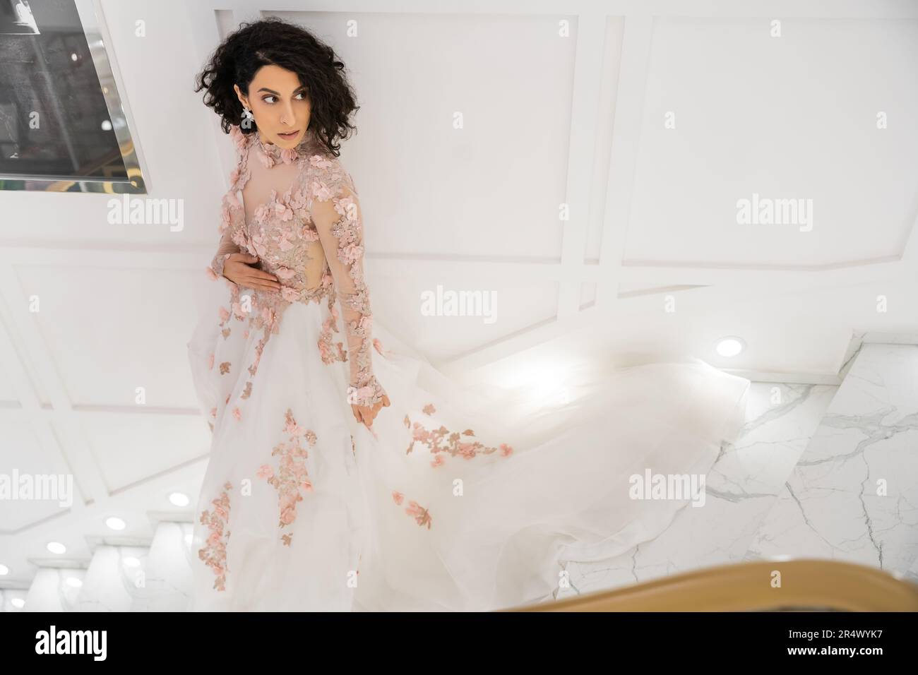 high angle view of brunette middle eastern woman with wavy hair standing in gorgeous and floral wedding dress with train while looking away in luxurio Stock Photo