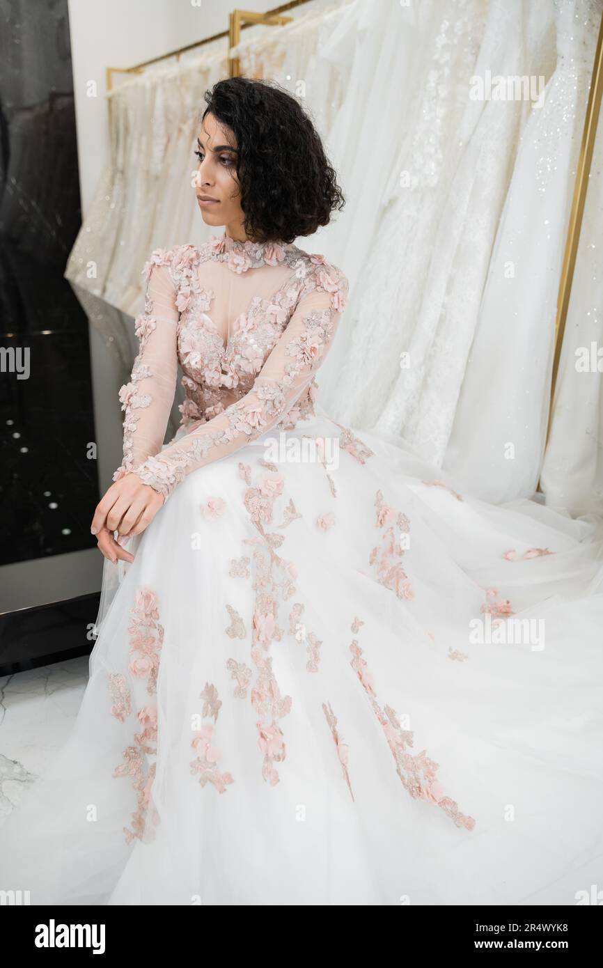 brunette middle eastern woman with wavy hair sitting in gorgeous and floral wedding dress near blurred and white gown inside of luxurious bridal salon Stock Photo