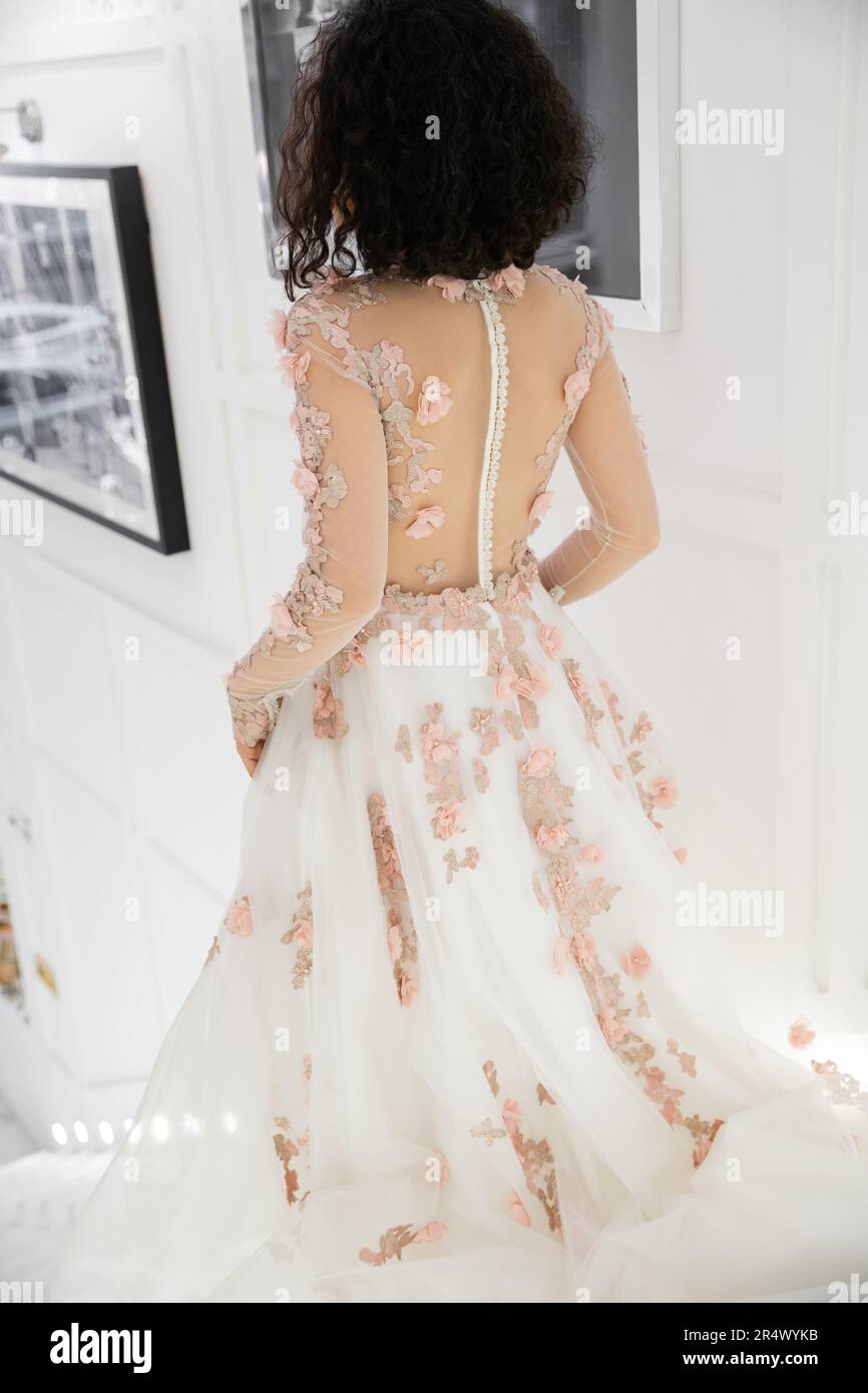 back view of brunette middle eastern woman with wavy hair walking in floral wedding dress inside of luxurious bridal salon, charming and elegant bride Stock Photo