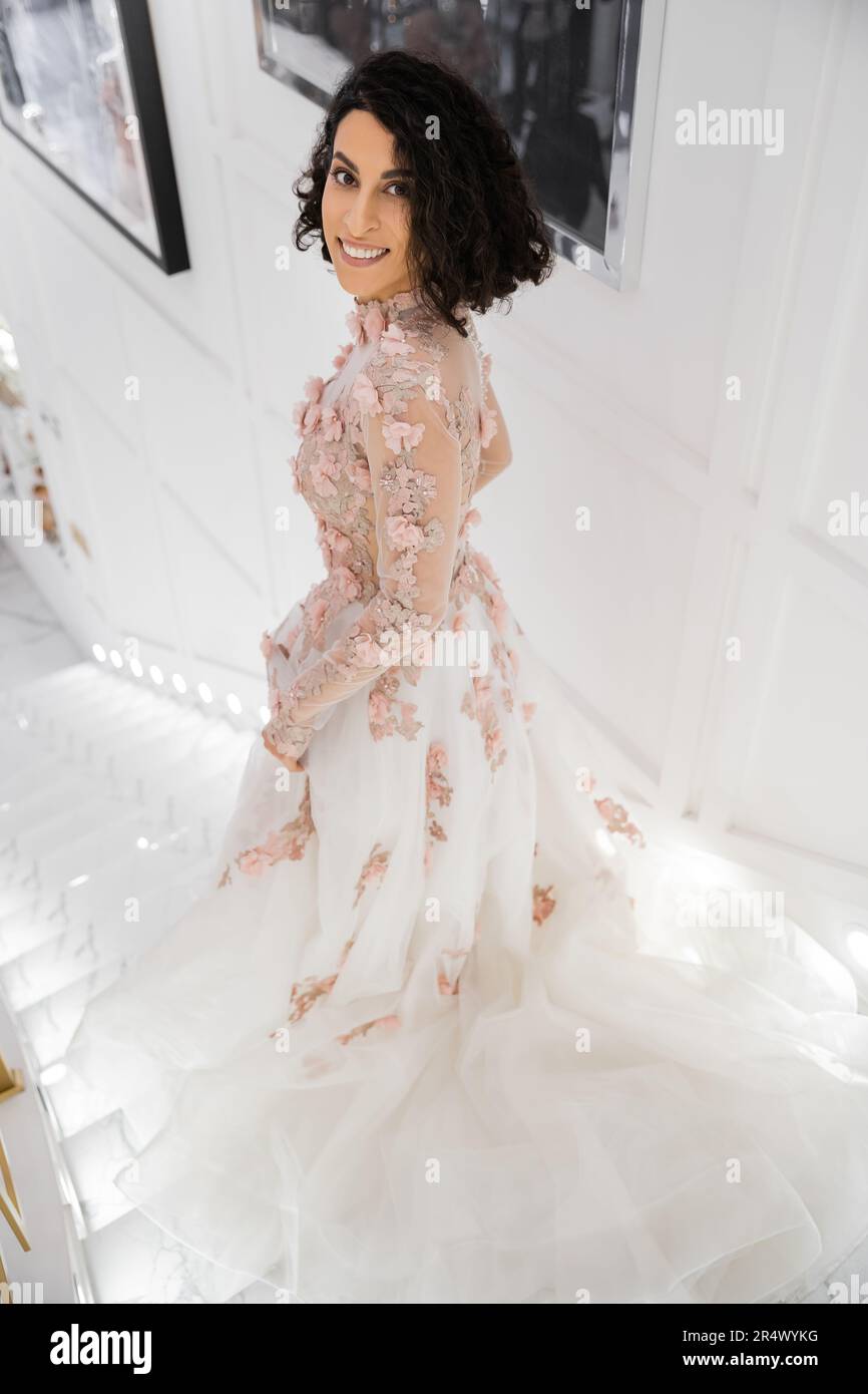 brunette middle eastern woman with wavy hair standing in gorgeous and floral wedding dress while smiling in luxurious bridal salon and looking at came Stock Photo