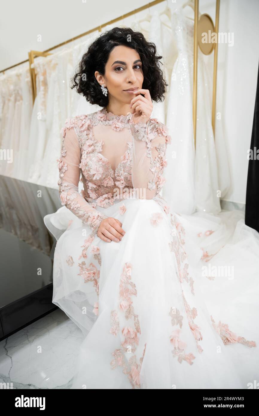 brunette middle eastern woman with wavy hair sitting in gorgeous and floral wedding dress near blurred and white gown inside of luxurious bridal salon Stock Photo