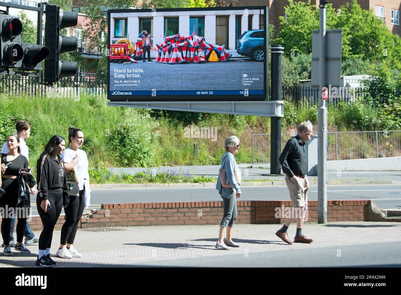 pedestrians wait at a crossing in kingston, surrey, england, in front of a digital billboard from the dvla warning of avoiding vehicle tax Stock Photo