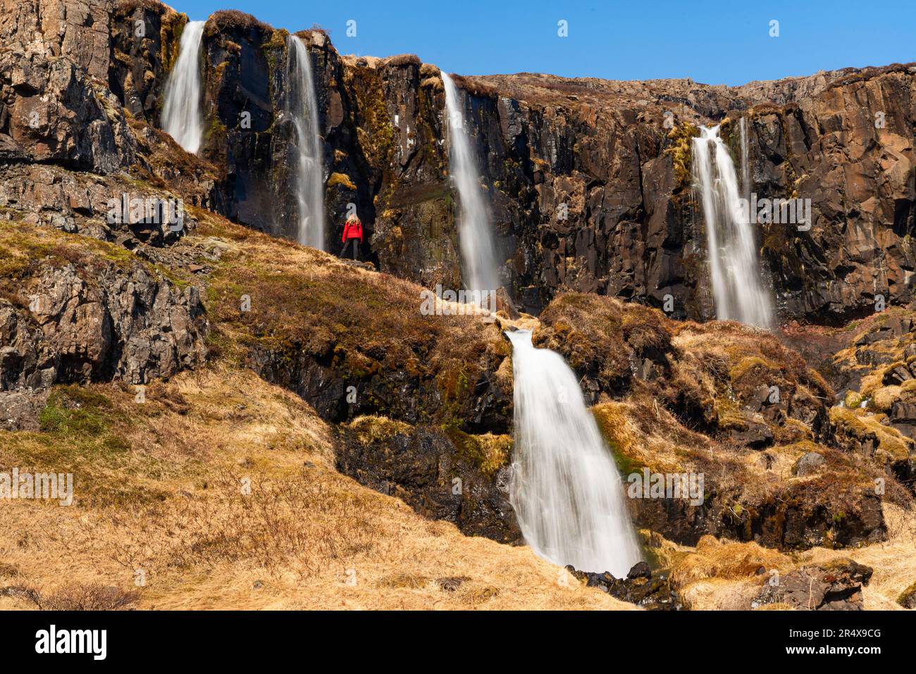 Scenic view of a woman standing on a cliff side slope in front of a series of waterfalls, some above and below, flowing from the craggy cliffs of t... Stock Photo
