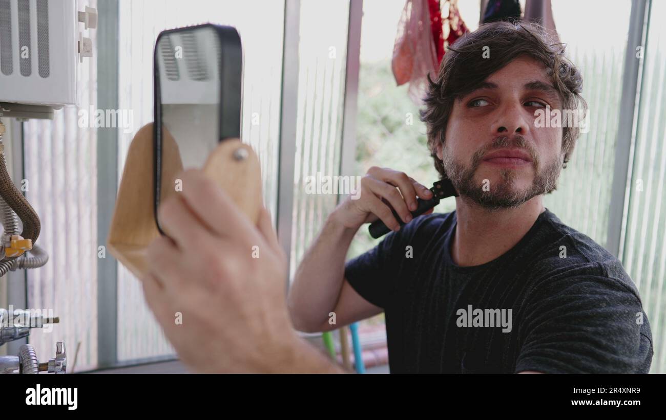 One young man shaving beard with trimmer on the go while standing in apartment balcony and holding a mirror Stock Photo