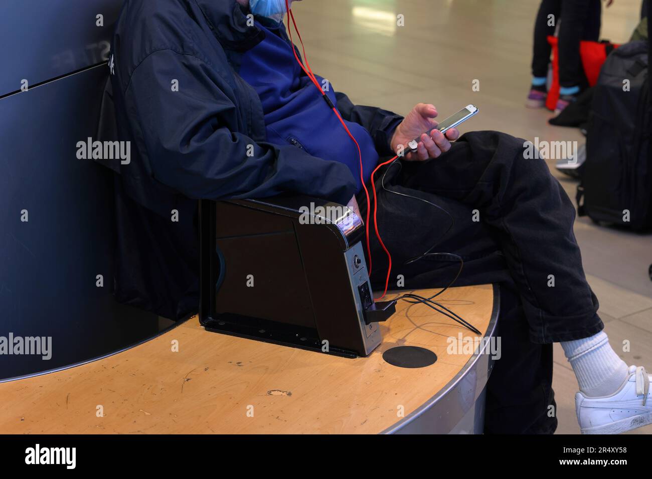 A person simultaneously listens to music while charging a smartphone from an electrical outlet in a public waiting area. Stock Photo