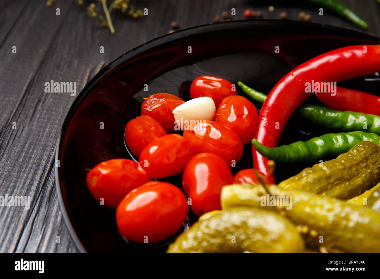 Canned cherry tomatoes, cucumbers, chili peppers in a round black plate on a dark background, close-up. Mix of pickled vegetables, spices Stock Photo