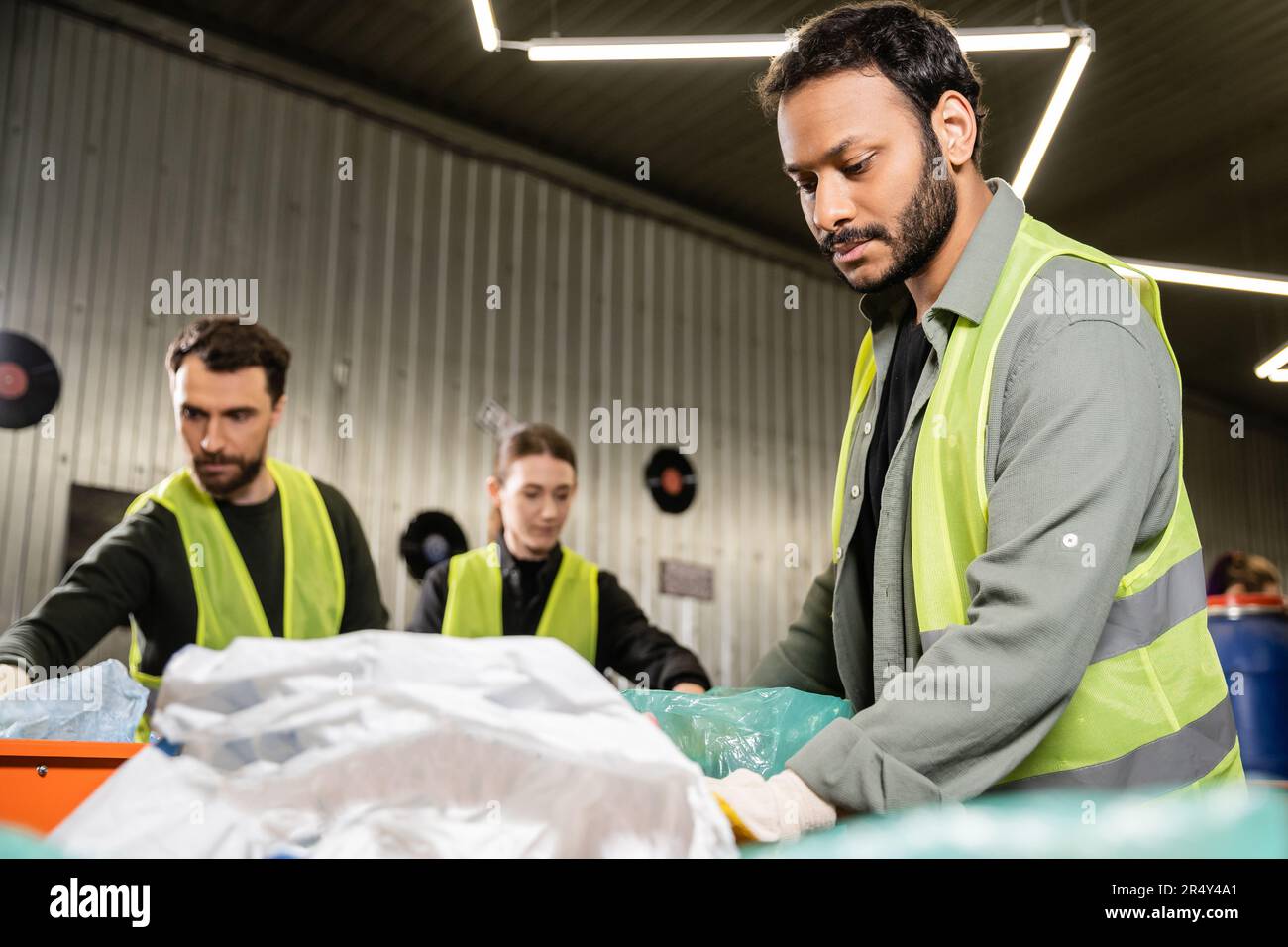 Indian worker in glove and protective uniform standing near plastic bag while separating trash near blurred conveyor and colleagues at background in w Stock Photo