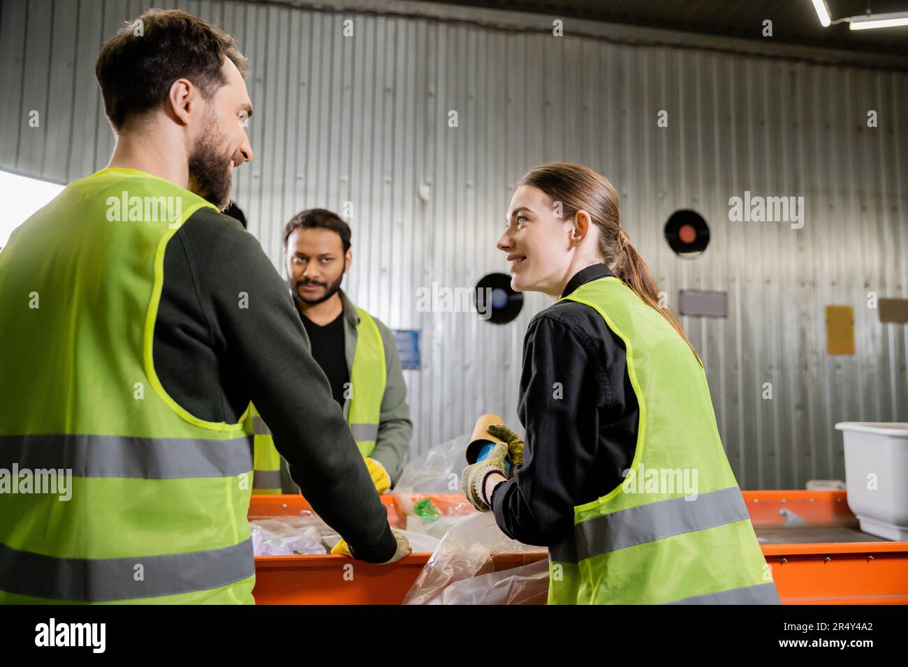 Smiling woman in protective uniform and gloves holding garbage and talking to interracial colleagues while working together near conveyor in waste dis Stock Photo