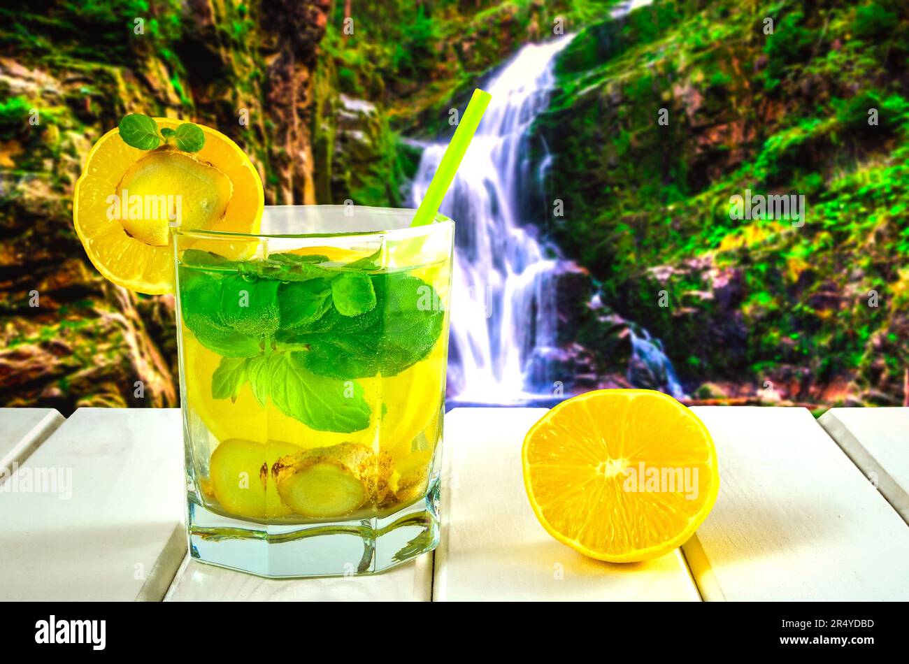 Drink in a glass on a white wooden table. Drink with lemon, ginger and mint with mountain waterfall in the background. Stock Photo