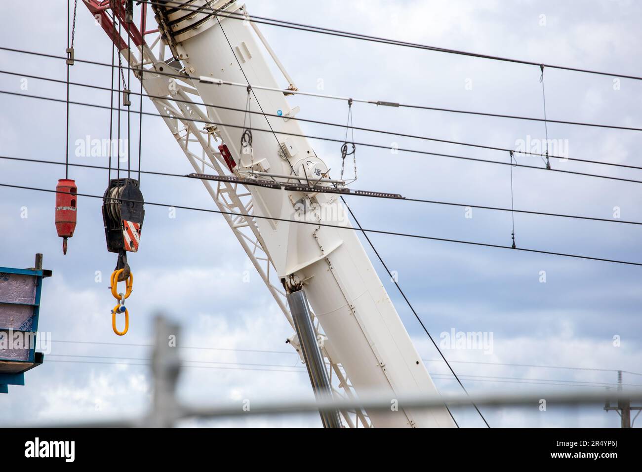 White crane boom with yellow hooks and train power lines.  Close up against a grey sky. Small section of Rail bridge. Stock Photo