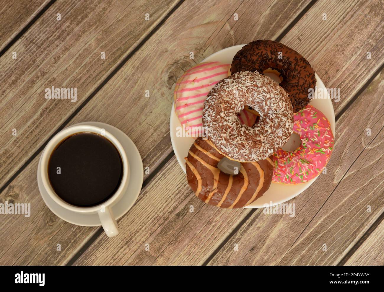 A cup of hot black coffee and a plate with several fresh donuts in multi-colored glaze on a light wooden table. Top view, flat lay. Stock Photo