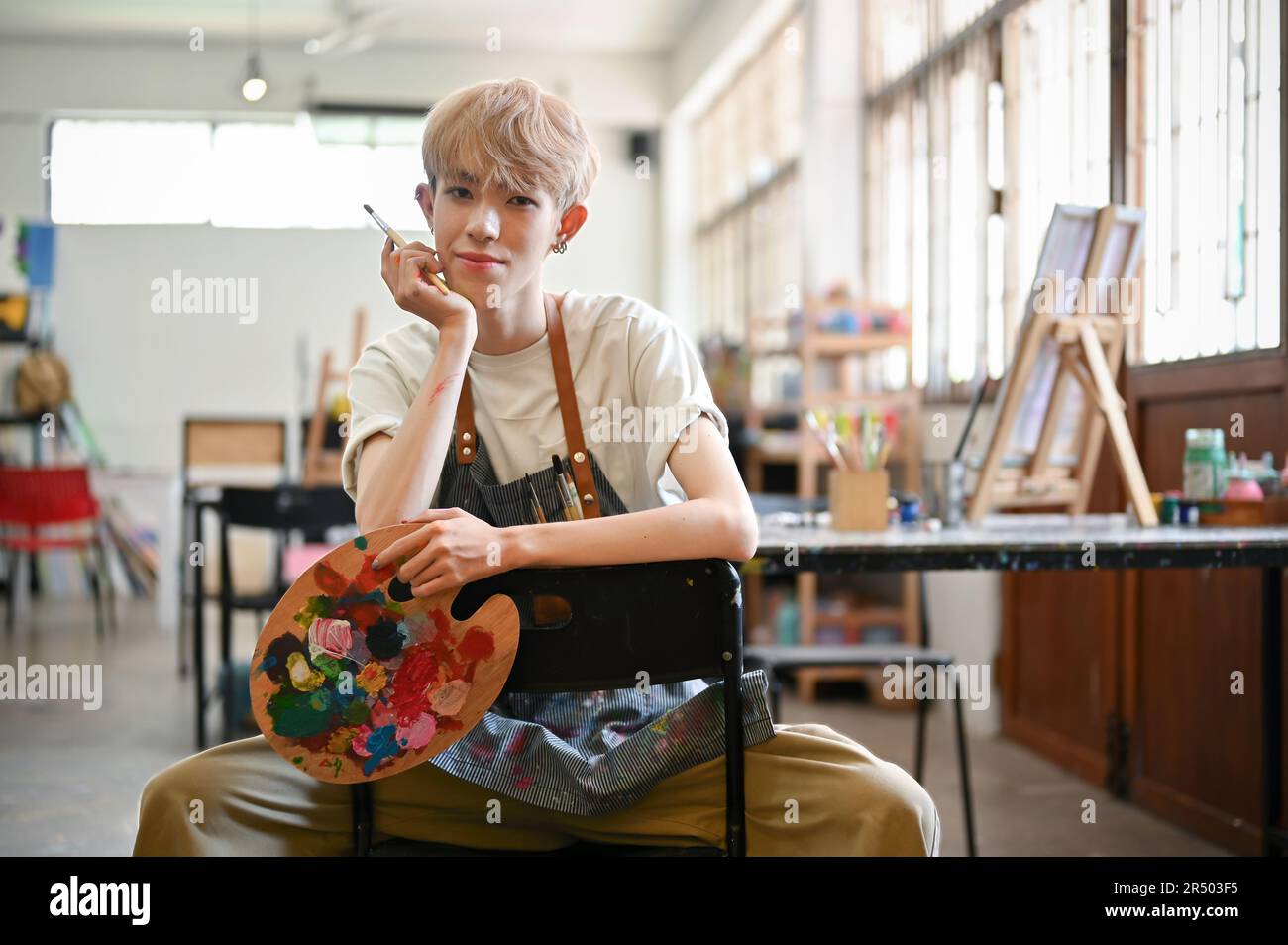 Portrait of a talented and creative gay man or queer artist in an apron is sitting on a chair in his creative studio with a paintbrush and art palette Stock Photo