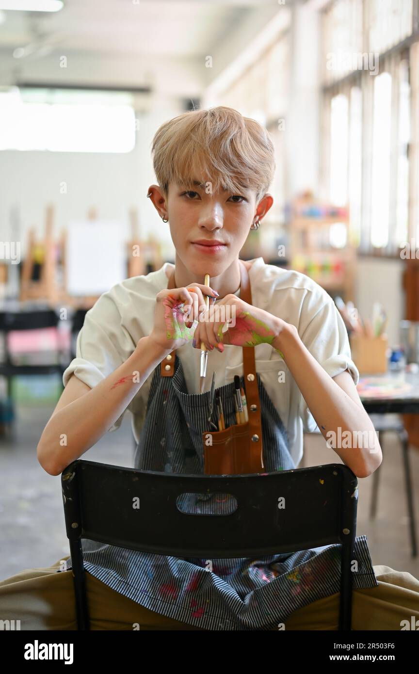 A portrait of an attractive and confident young Asian queer or gay man artist in an apron sitting on a chair in his creative studio. LGBTQ+ and creati Stock Photo