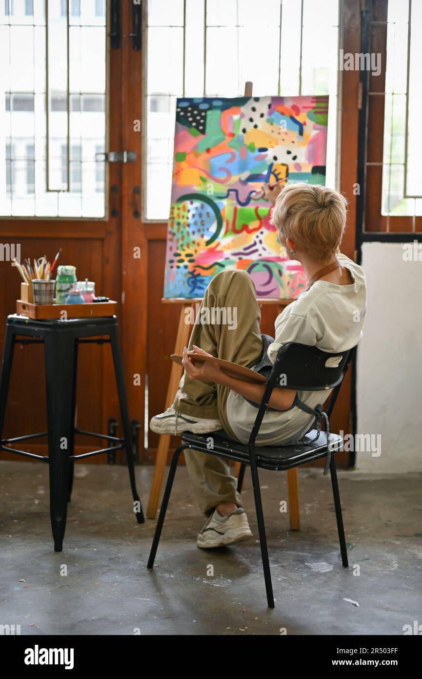 Back view. A talented young Asian queer artist sits on a chair and ...