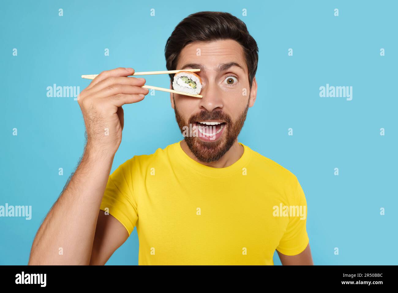 Funny man hiding his eye with tasty sushi roll on light blue background
