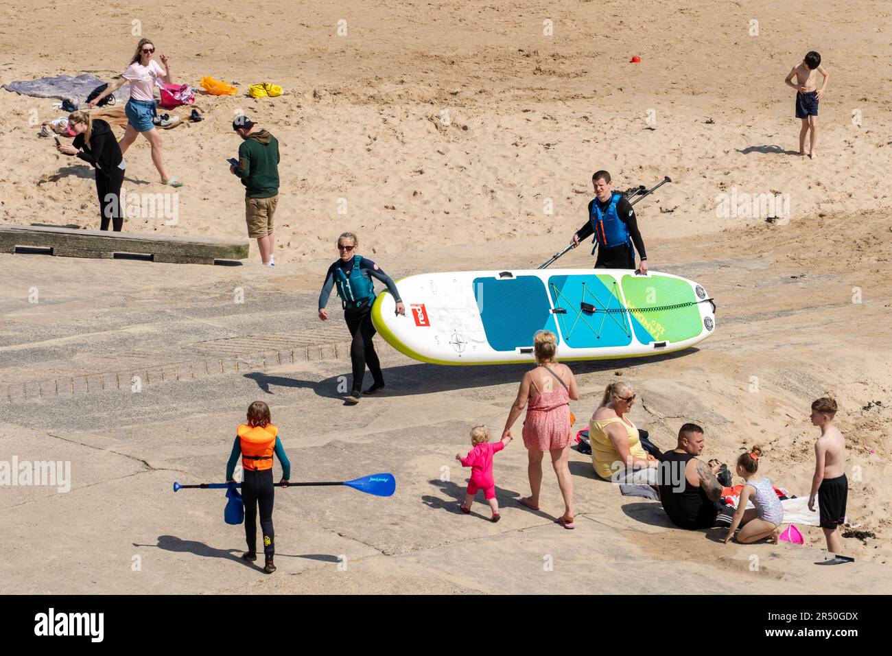 People enjoying the beach in sunny weather, including stand up paddleboarders with their board. Cullercoats Bay, North Tyneside, UK Stock Photo