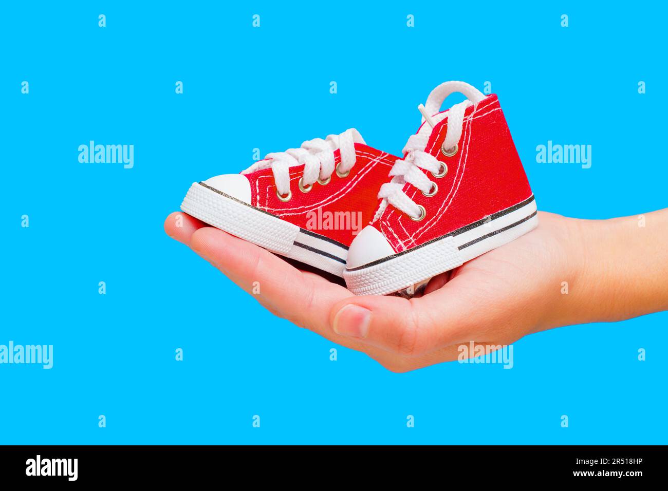 Pair of tiny red high-top canvas shoes in hand isolated on blue with copy space. Creative baby footwear concept. Stock Photo