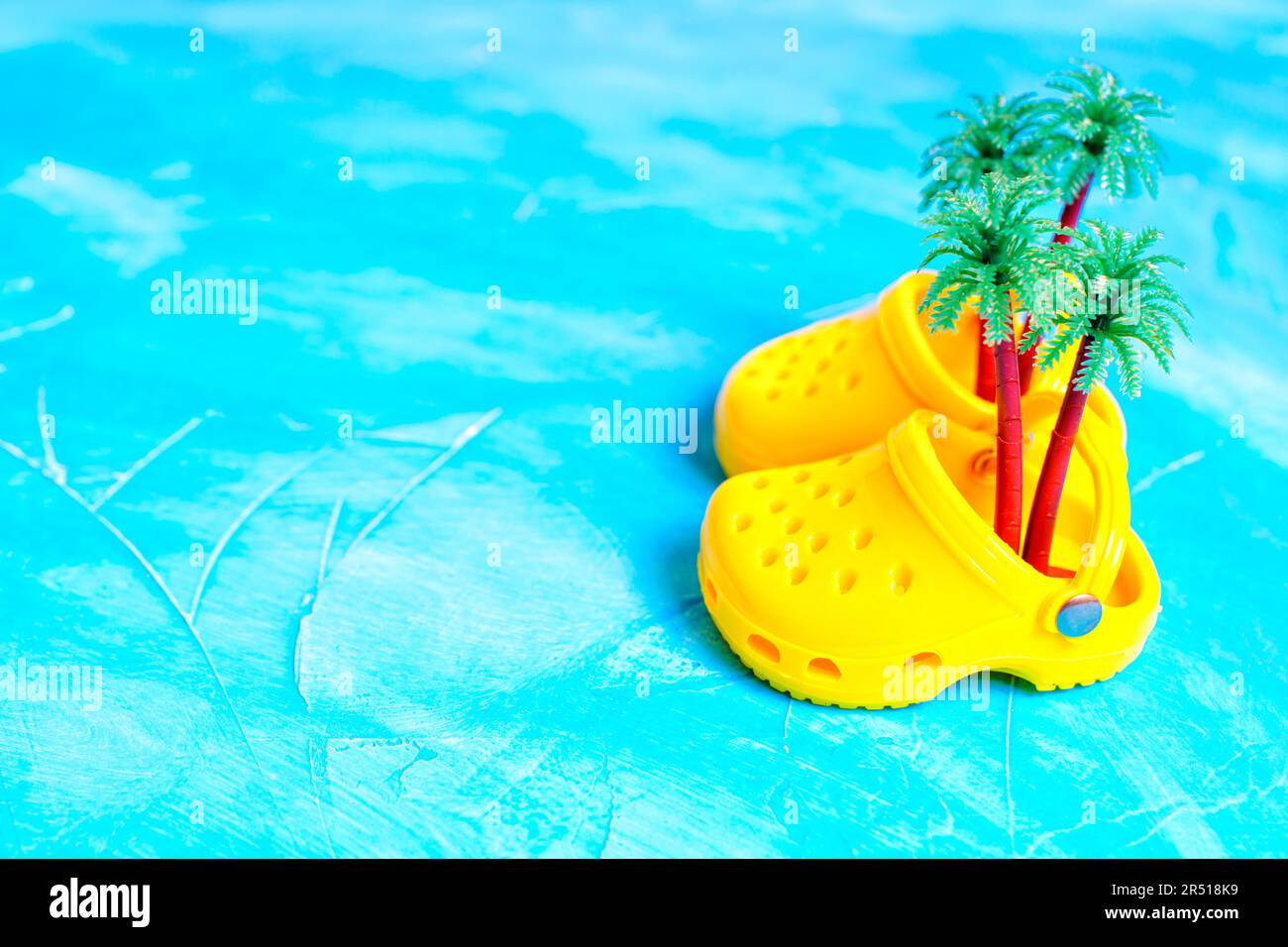 Tiny perforated yellow clogs with toy palm trees isolated on a blue background. Vivid beachwear promotions and summer footwear concept. Stock Photo