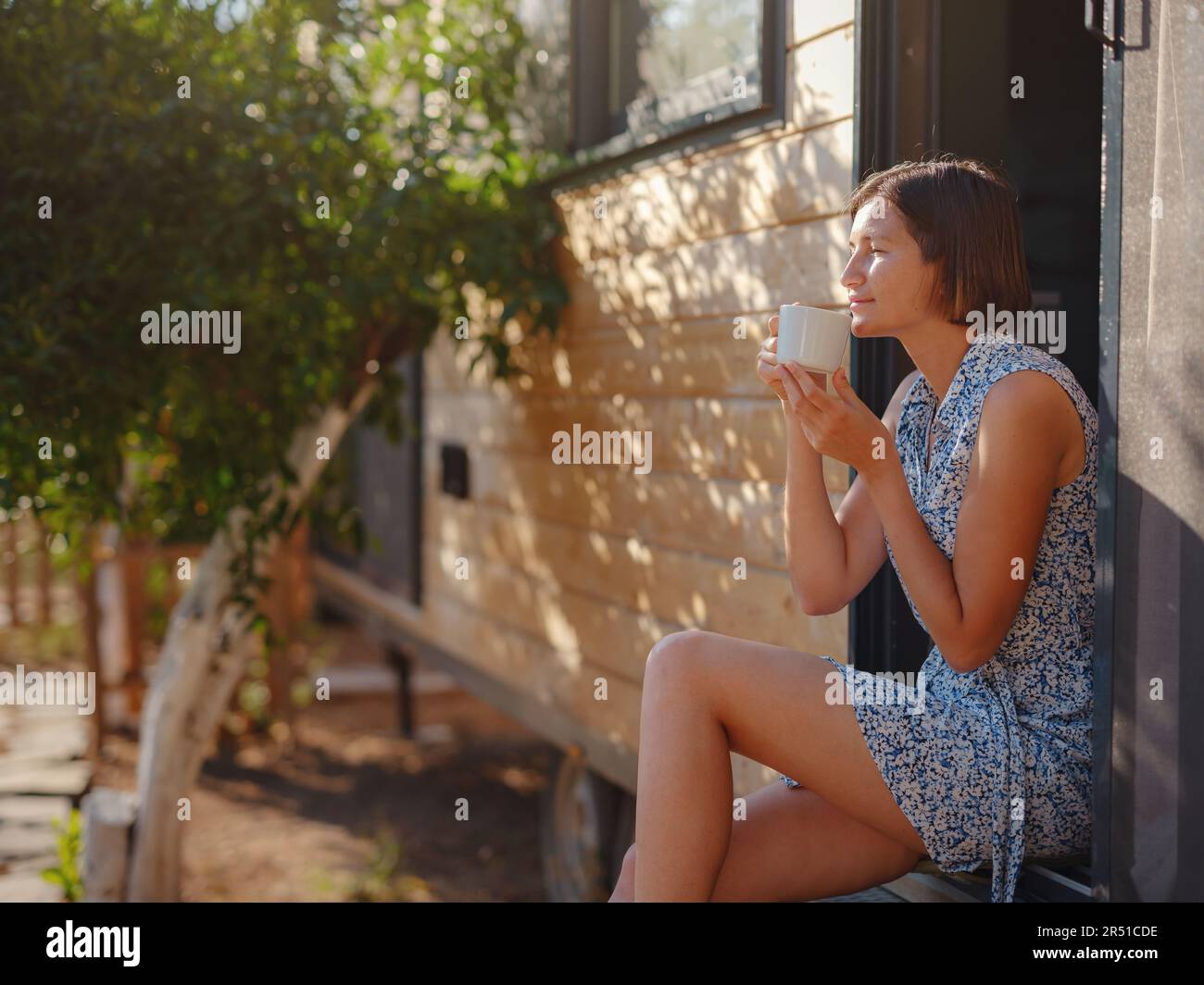 Woman is drinking coffee , morning routine near her Tiny house. First property. Small apartment in summer garden. Minimalism. Moving in. Living alone. Charming trailer house Stock Photo