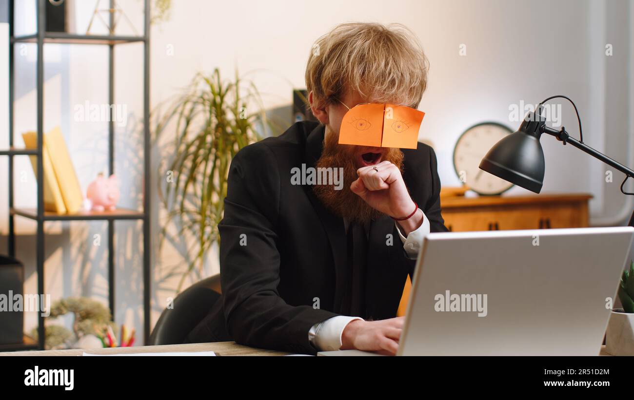 Young business man with fake eyes painted on paper stickers sleeping at  workplace in office Stock Photo