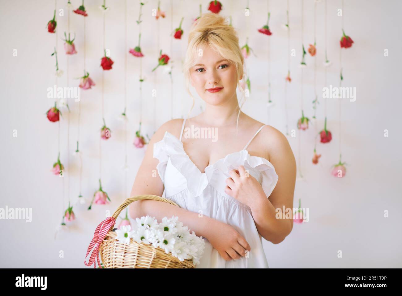 Studio portrait of pretty young teenage 15 - 16 year old girl wearing summer dress, posing on white background with hanging roses, holding basket with Stock Photo