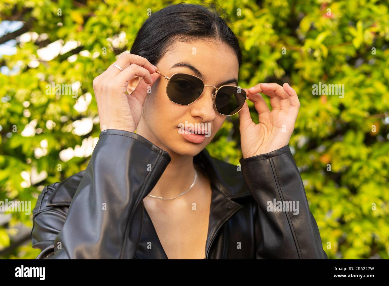 Portrait of confident young female with black hair in leather jacket putting on sunglasses while standing near green bushes Stock Photo