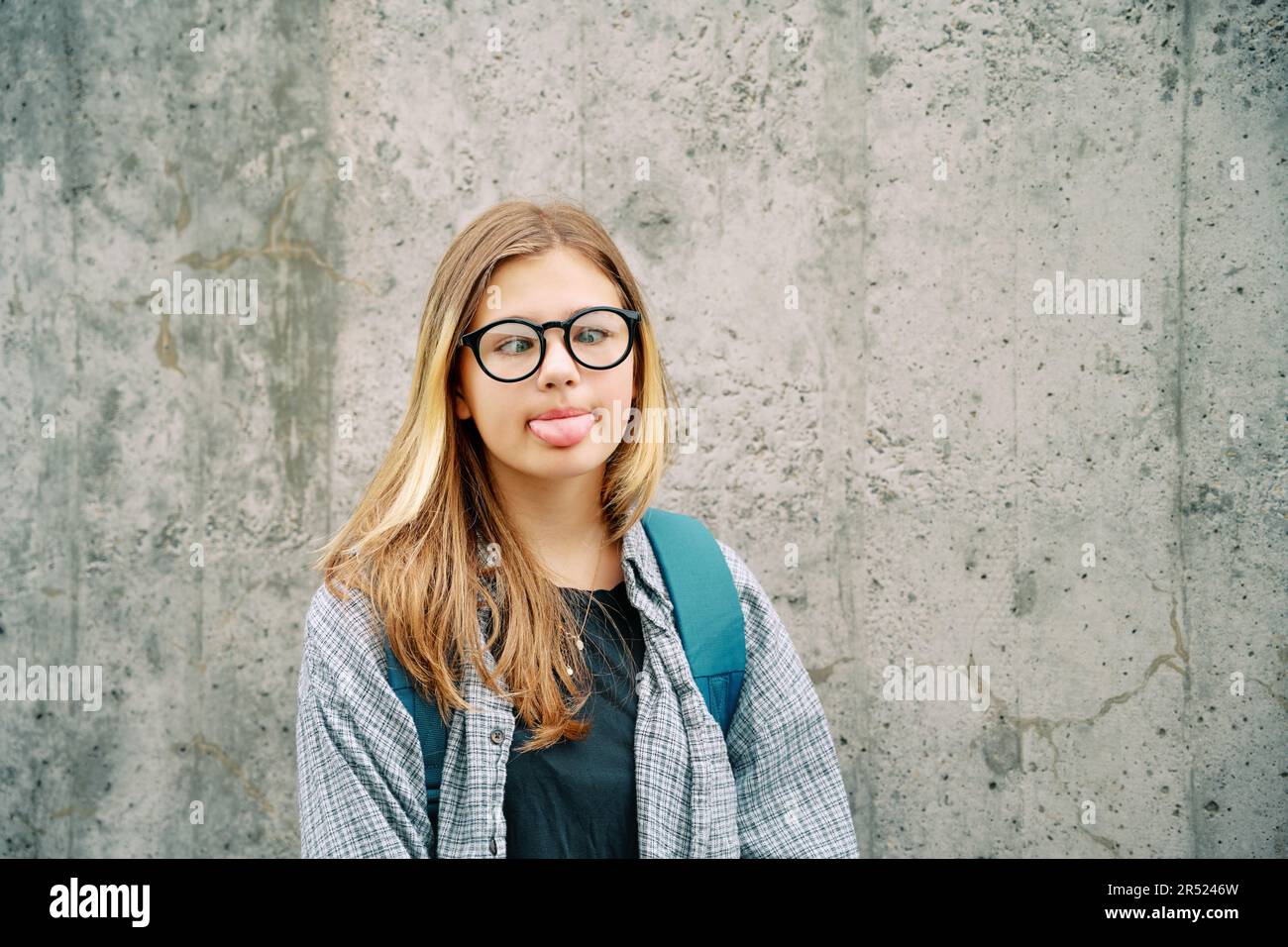 Outdoor portrait of silly young teenage kid girl pulling a tongue, crossing eyes over her nose, wearing glasses and backpack, posing on grey wall back Stock Photo
