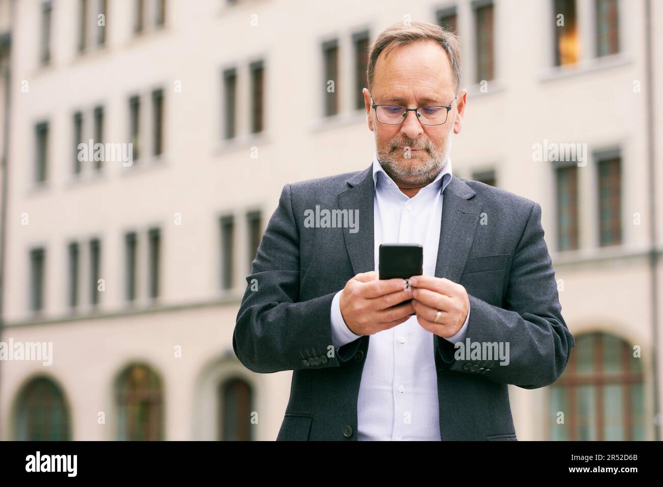 Outdoor portrait of middle age man, wearing glasses and grey suit, holding smartphone Stock Photo