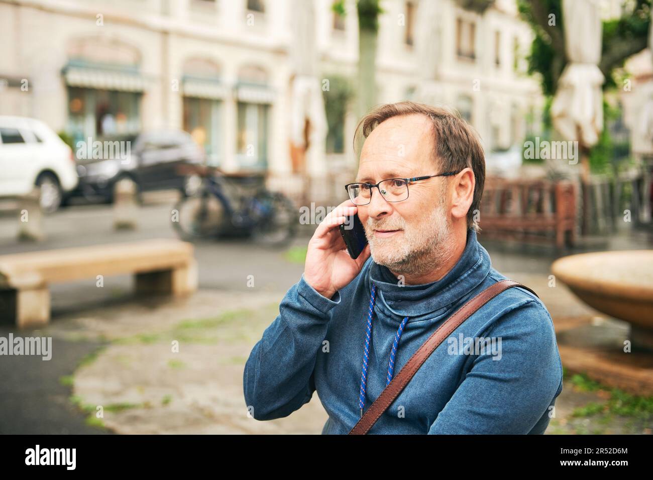 Outdoor portrait of middle age man talking on the phone, wearing glasses and blue sweatshirt, holding smartphone next to ear Stock Photo