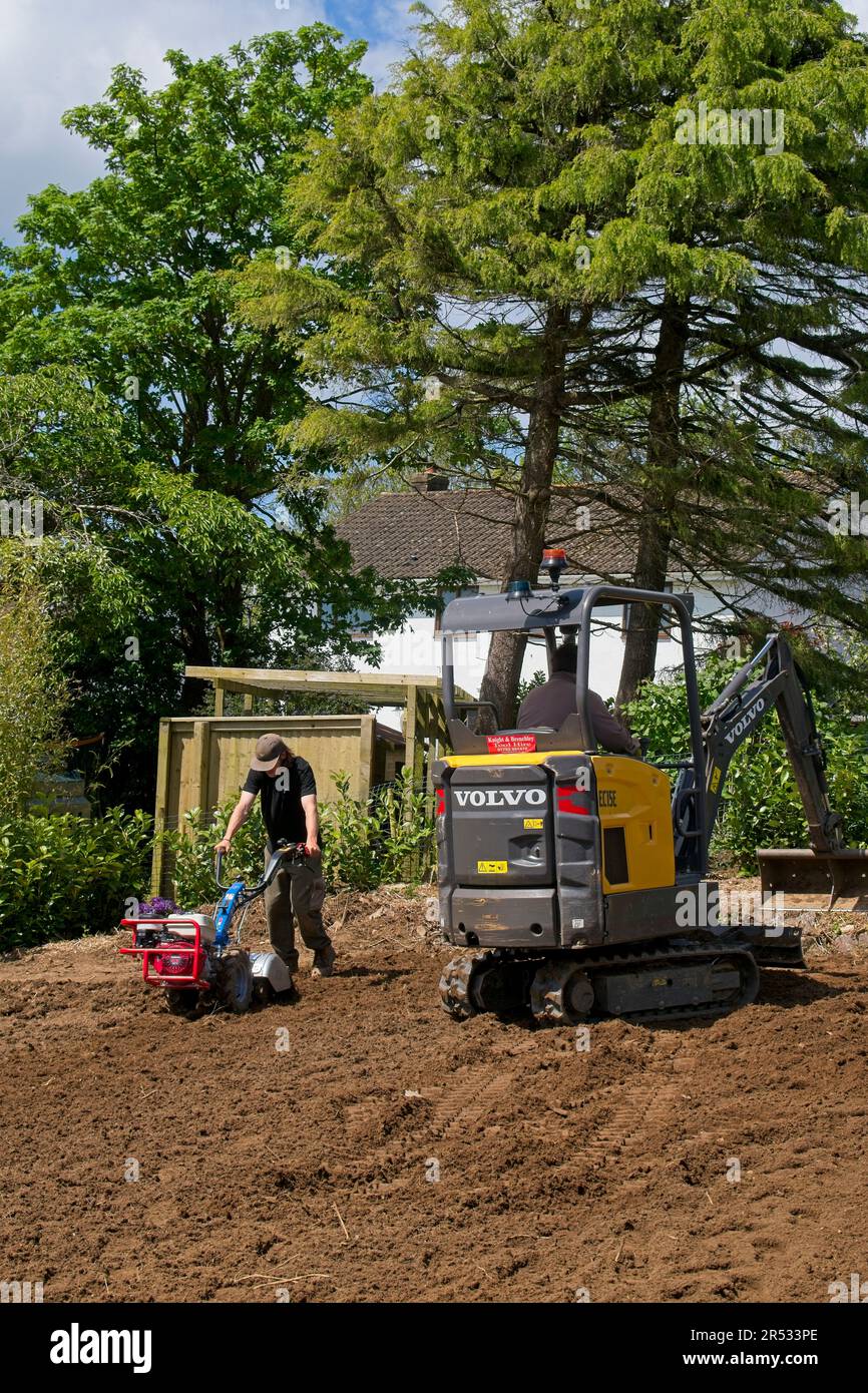Gower Wales UK May 2023 Machinery preparing ground for replacement grass lawn. Mechanical digger and rotavator in use. Trees and bushes in background. Stock Photo