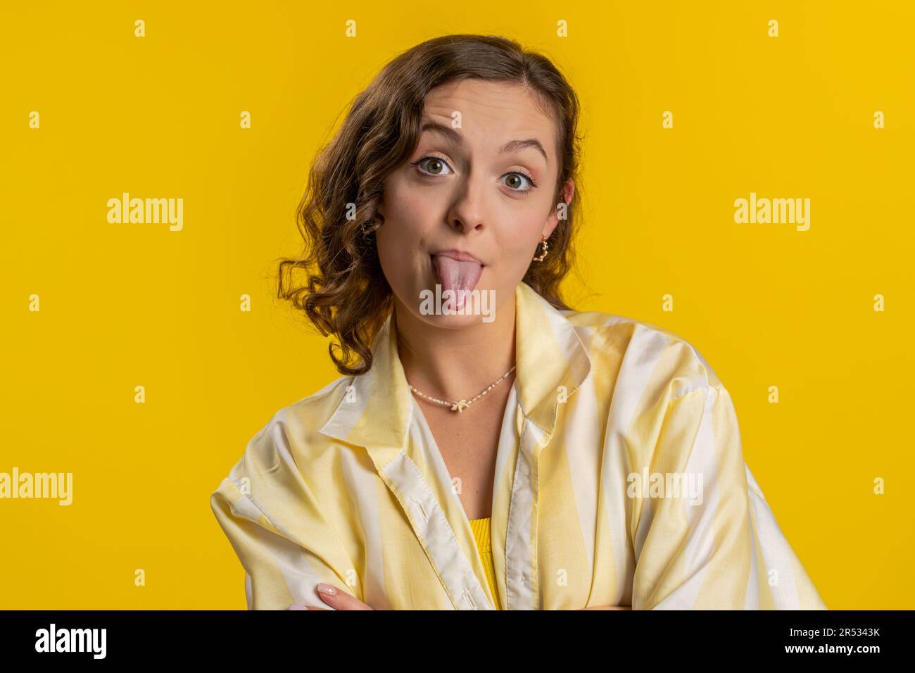 Cheerful funny young woman showing tongue making faces at camera, fooling around, joking, aping with silly face, teasing, bullying, abuse. Girl isolated alone on yellow studio background. Lifestyles Stock Photo