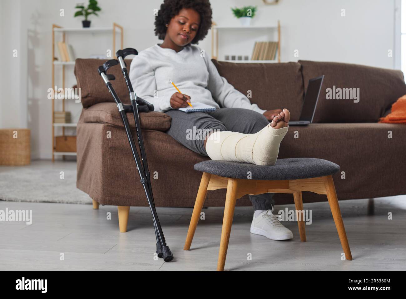 Beautiful, sweet african american woman works at home in the living room with laptop and makes notes in notebook, sitting on the sofa with broken Stock Photo
