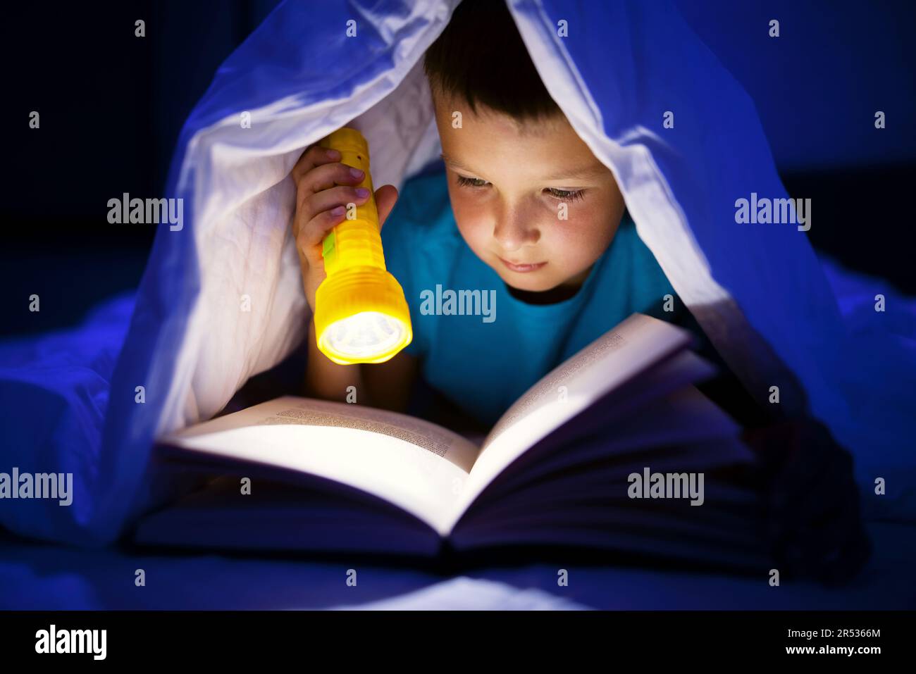 A young boy reading a book under the covers with a flashlight at dark