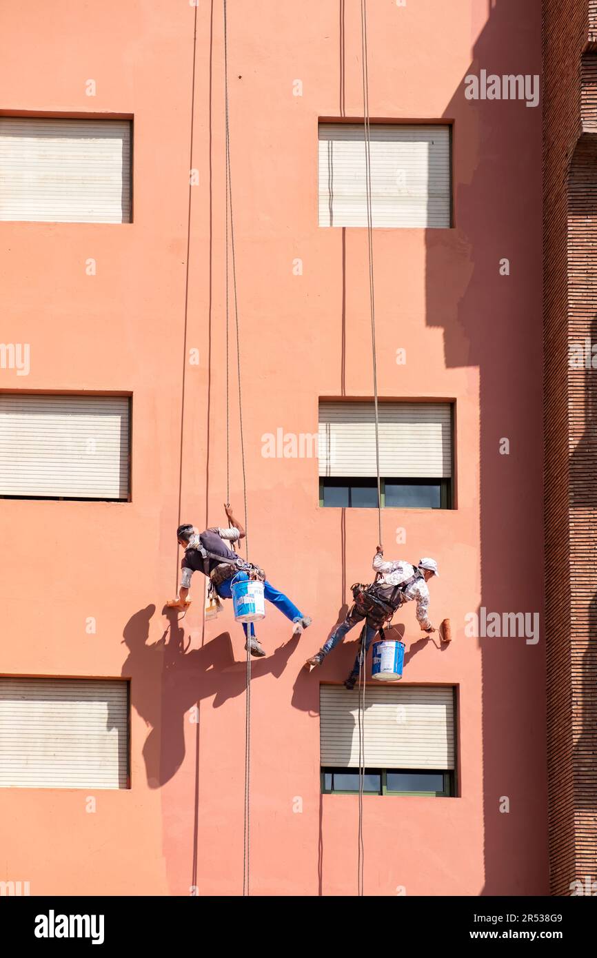 Two rope access painters abseiling down the wall of a building giving the walls a fresh coat of paint, using paint rollers, as they move down the wall Stock Photo