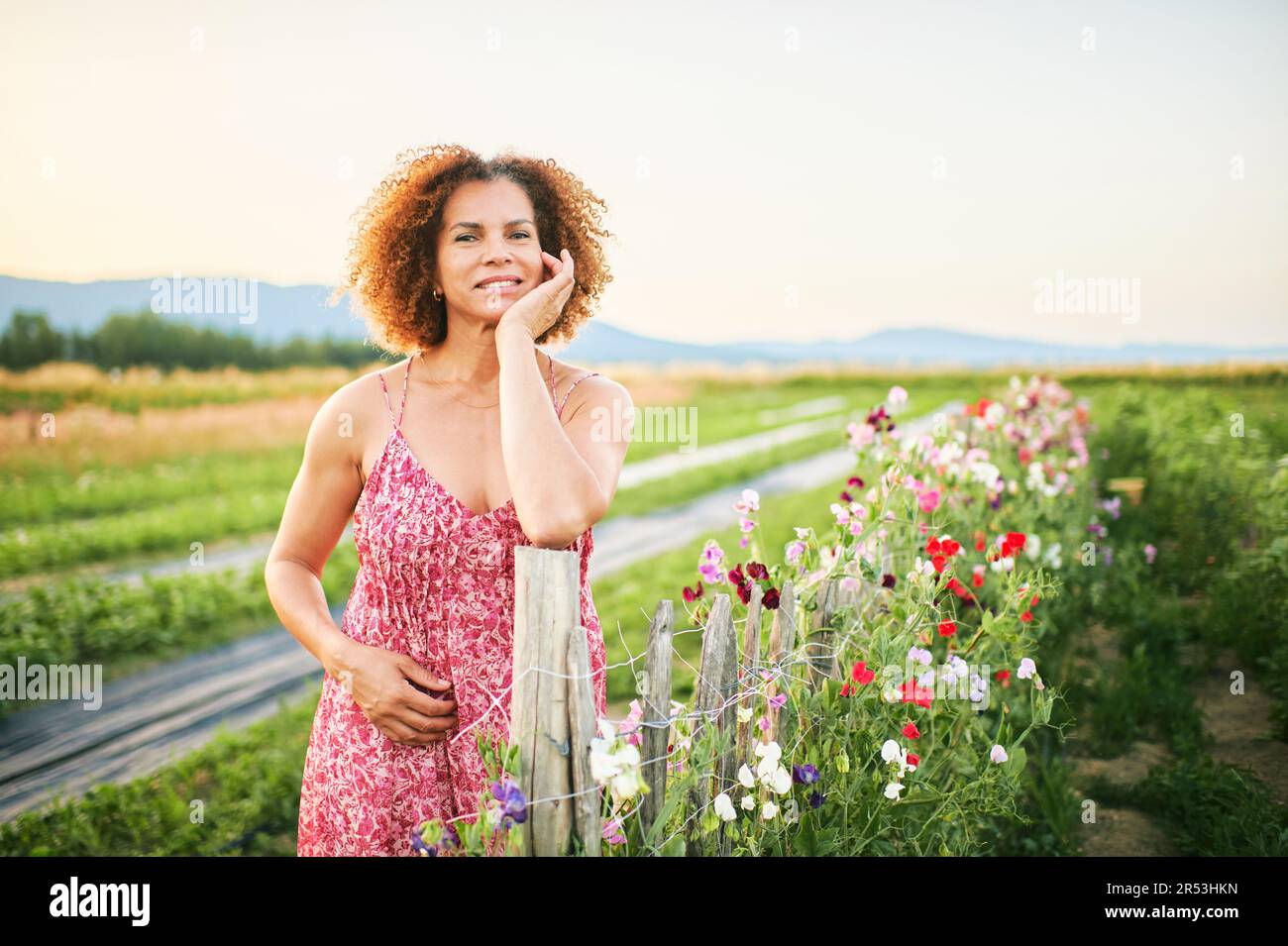 Outdoor portrait of beautiful middle age woman enjoying nice suuny evening in countryside, leaning on flower fence, wearing red dress Stock Photo