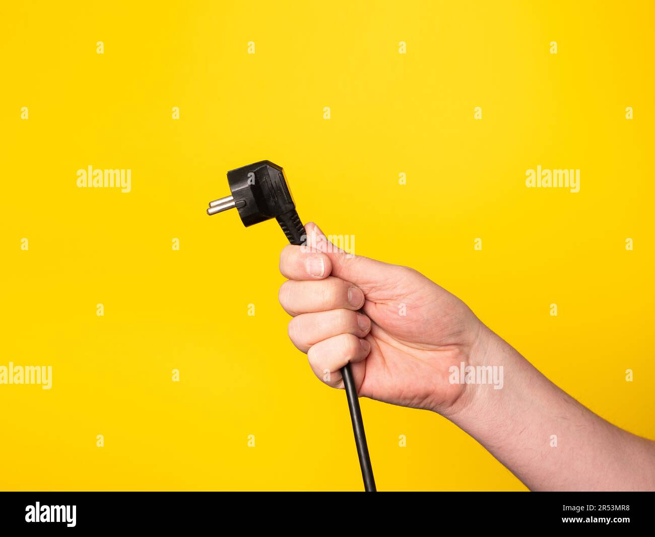 A hand holds a plug to which a black cable is attached.  No face, yellow background. Stock Photo