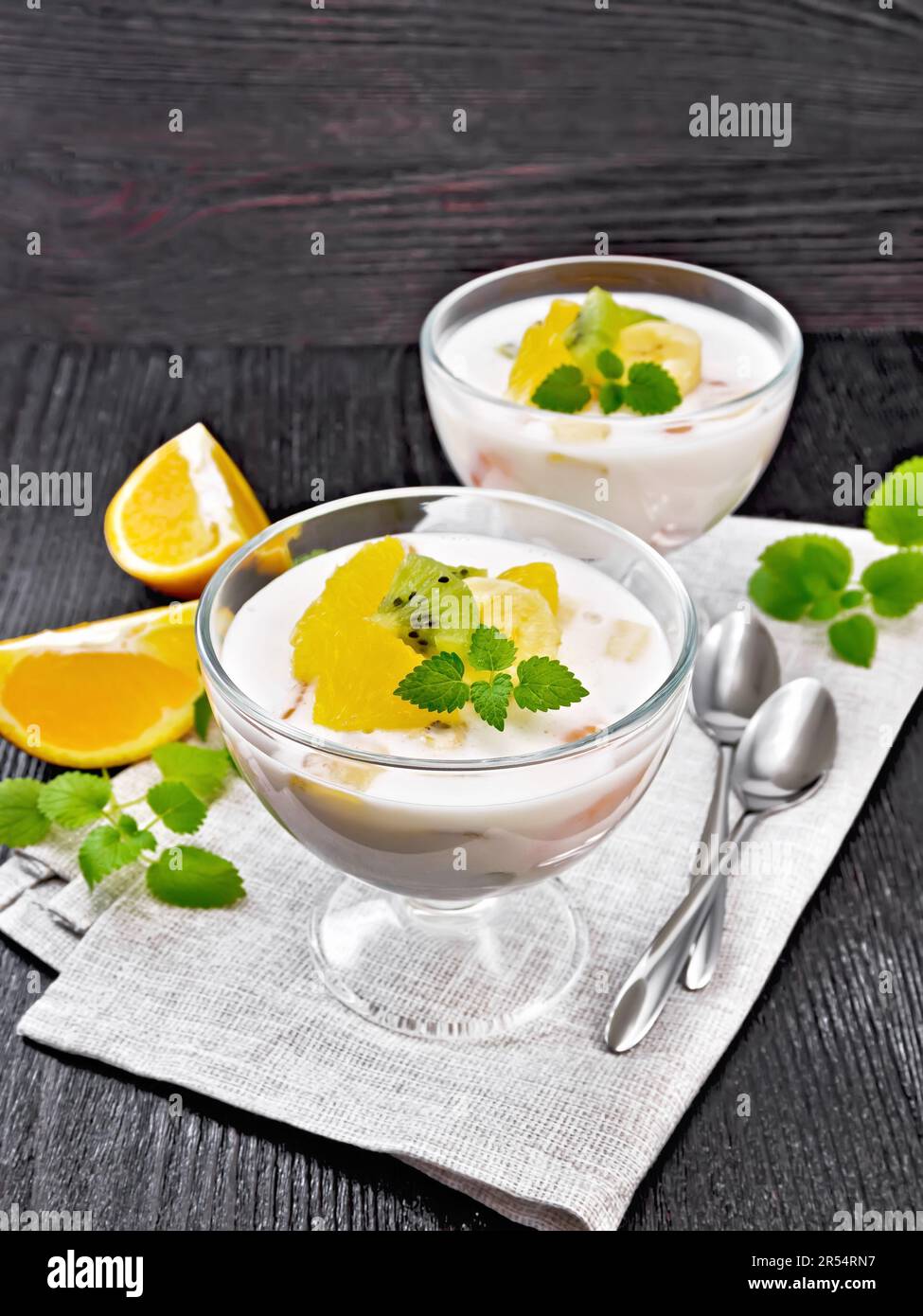 Dessert airy yogurt with orange, banana and kiwi in two glass bowls, fruits, spoons and mint on a napkin against dark wooden board Stock Photo