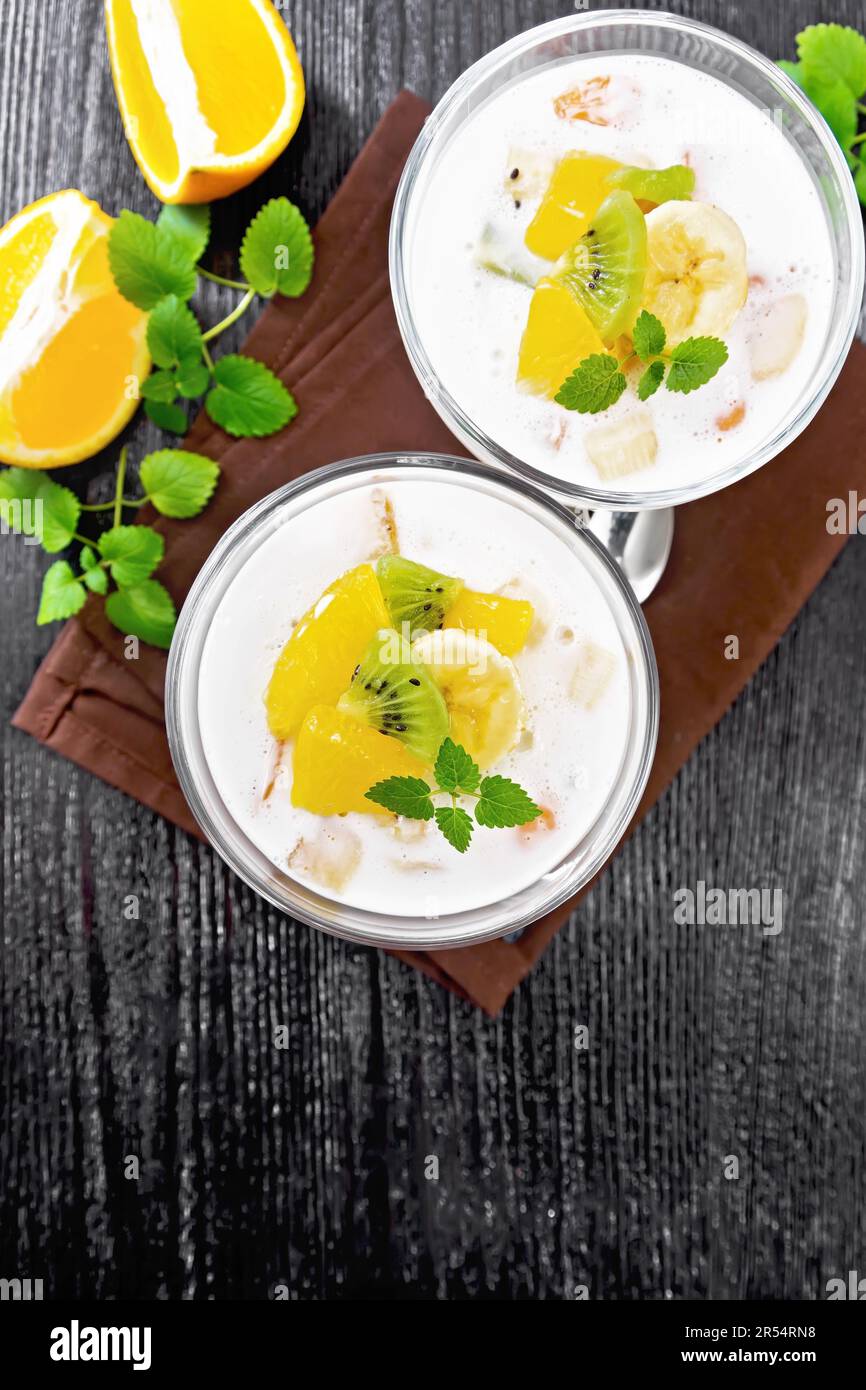 Dessert airy yogurt with orange, banana and kiwi in two glass bowls, fruits, spoons and mint on a brown towel against a background of wooden board fro Stock Photo