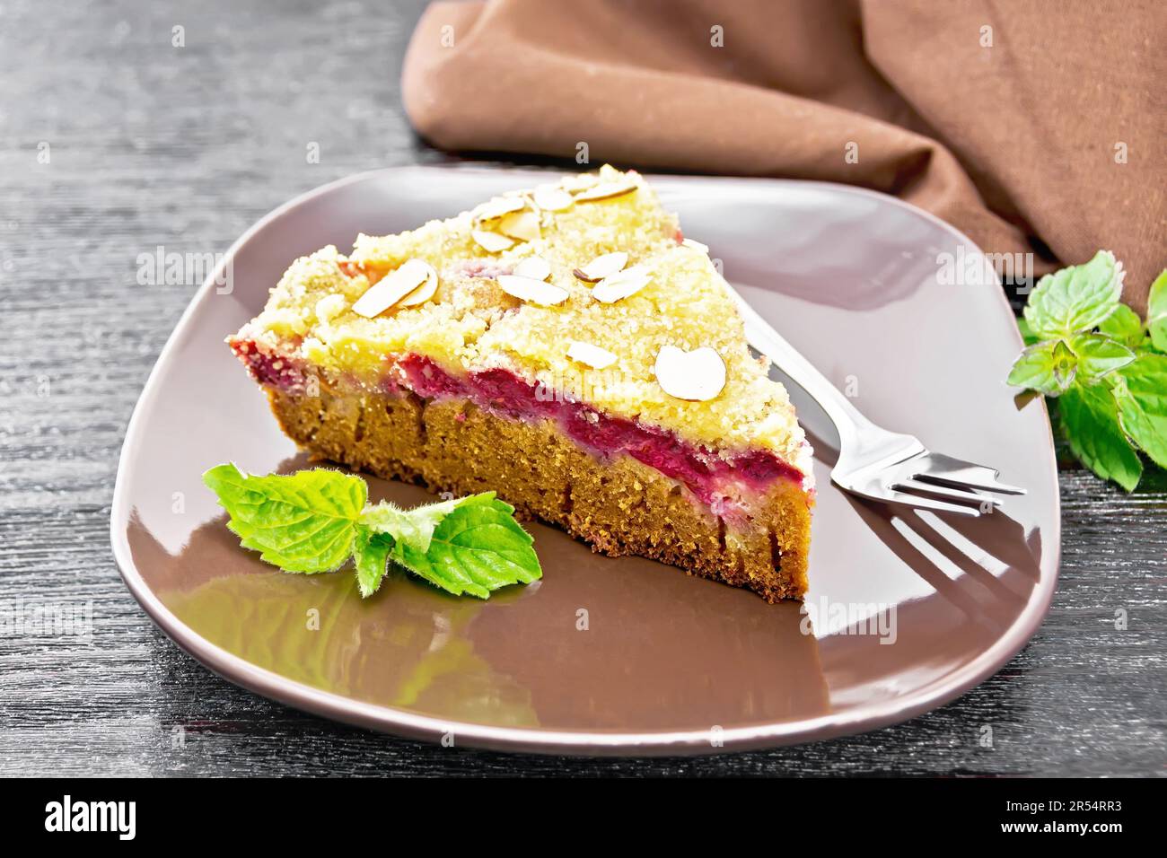 A piece of sweet cake with raspberries, butter crumble and almond flakes in a plate, mint and napkin on dark wooden board background Stock Photo