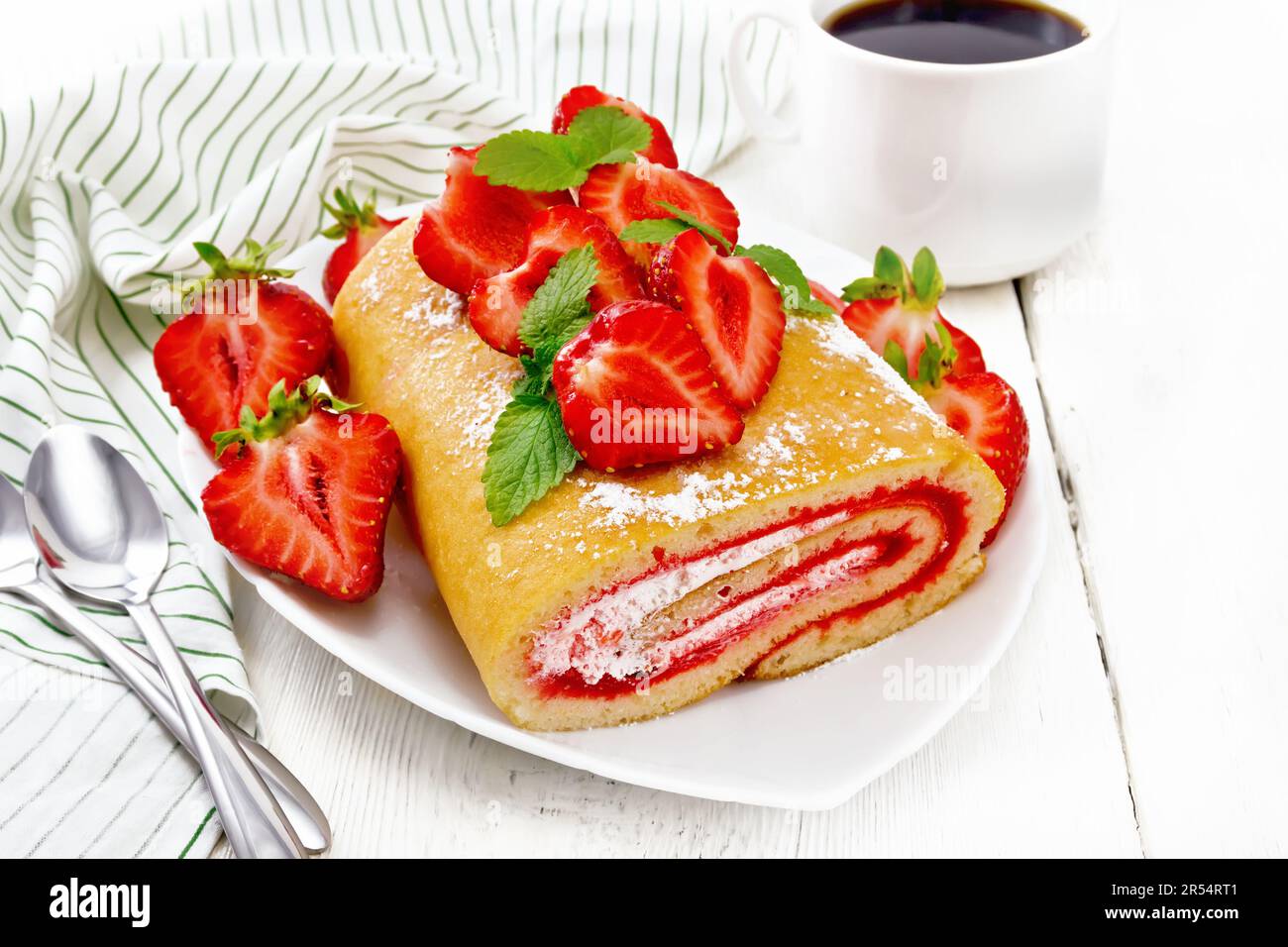 Roll with cream and strawberry jam, fresh strawberries and mint in a plate, napkin and spoons on white wooden board background Stock Photo