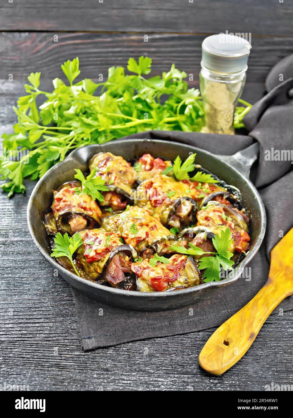 Baked eggplant rolls with meat, tomatoes and cheese in a frying pan, towel, parsley and spatula on a dark wooden board background Stock Photo
