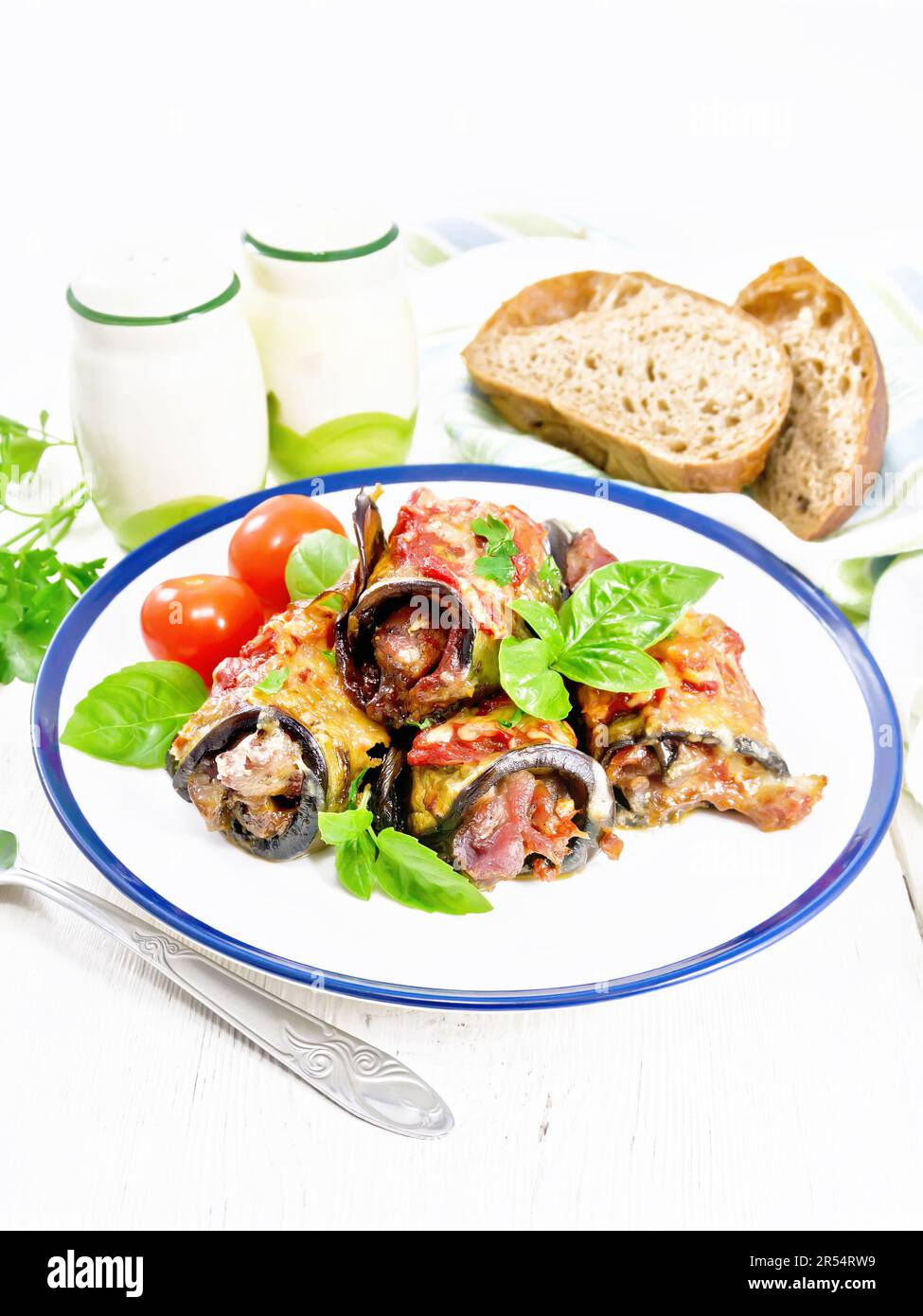 Baked eggplant rolls with meat, tomatoes and cheese in a plate, napkin, basil, bread and fork on a wooden board background Stock Photo
