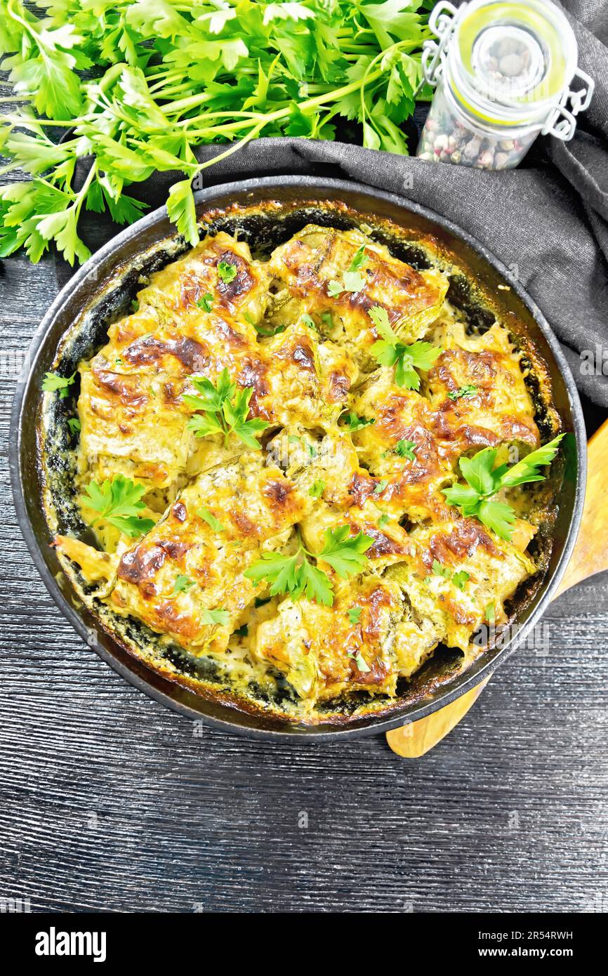 Baked zucchini rolls with meat, mayonnaise and cheese in a frying pan, napkin, parsley and spatula on a dark wooden board background from above Stock Photo