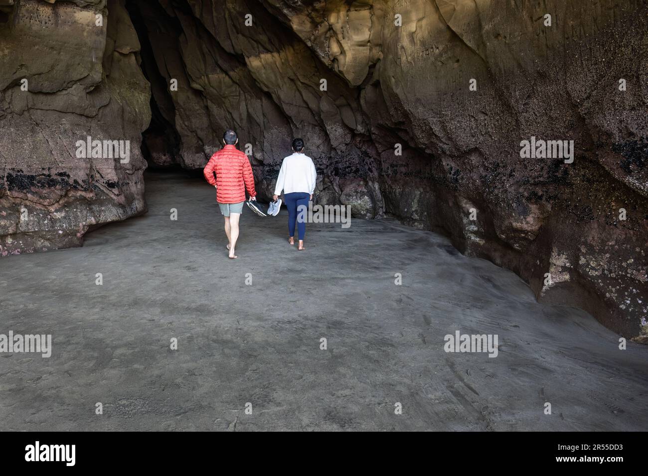 Young couple holding shoes in hands and walking barefoot towards a cave at Muriwai beach. Auckland. Stock Photo